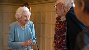 En las foto, la reina Isabel II sonriendo, vestida con un traje azul claro y adornada con un collar de perlas y un bastón en la mano.